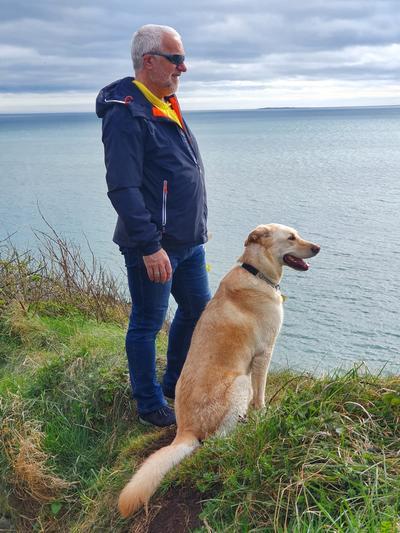 Tony standing on a cliff, looking out to sea with a golden Labrador dog